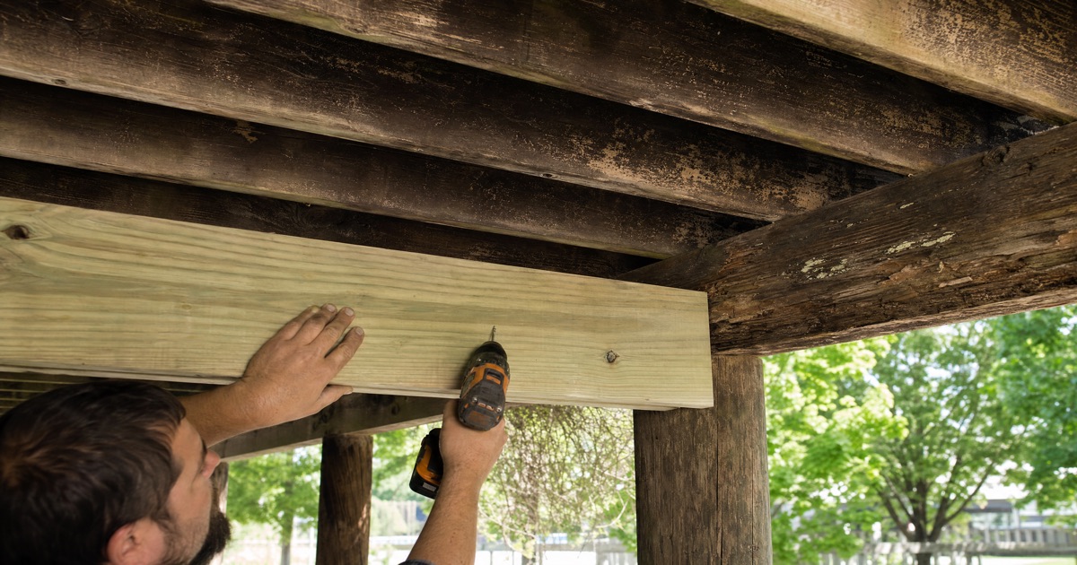Carpenter hands sistering a new pressure-treated wood joist alongside an old deteriorated wood joist under a backyard deck