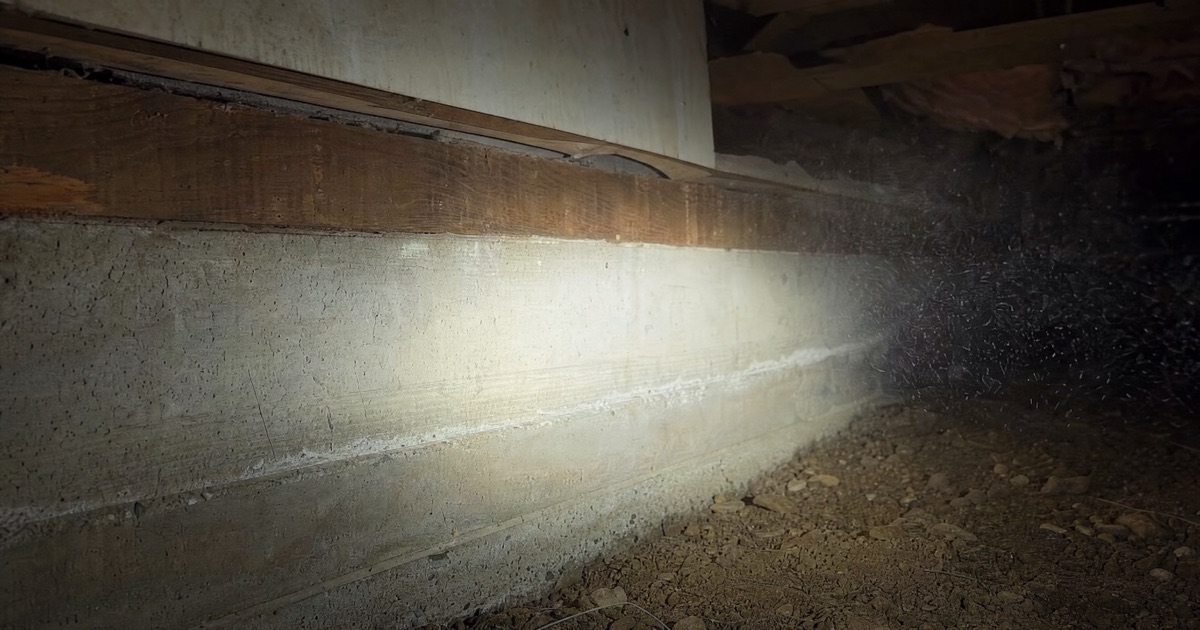 Crawl space view showing a wood sill plate resting on a concrete foundation wall, flashlight beam cutting through dusty air